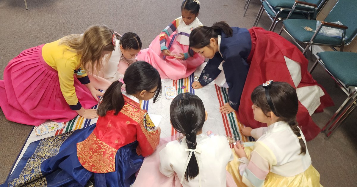 Students in hanbok kneeling around a striped mat playing gonggi at the Daegu rest stop.