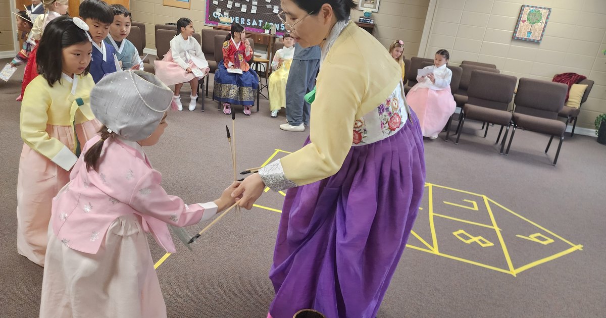 A teacher in hanbok handing throwing sticks to a student at the Daejeon rest stop, with the sabangchigi squares marked on the floor.